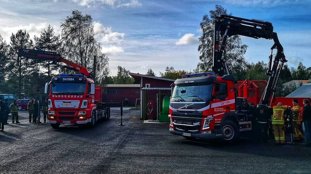 Kaksi nosturilla varustettua pelastuslaitoksen ajoneuvoa paloaseman pihalla, taustalla ihmisiä ja punaisia rakennuksia.